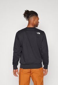 Schwarzer Pullover mit rundem Halsausschnitt und gerippten Bündchen. Verfügt über ein kleines weißes Logo auf der oberen Rückseite. Getragen mit orangefarbenen Hosen.