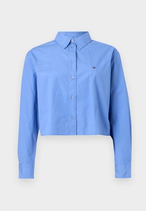 Innskåret blå skjorte med knapper, spiss krage, lange ermer og en liten brodert logo i rødt og hvitt på brystet.