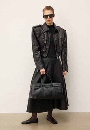 Zwarte leren jas met schouderepauletten, zwarte jurk en handtas. Zonnebril en zwarte platte schoenen maken de monochrome look compleet.