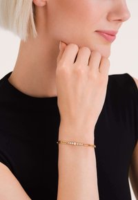 Zlatna narukvica s nizom malih prozirnih kamenčića, povezanim produljenim lančanim segmentima. Elegantni, minimalistički dizajn na zapešću.