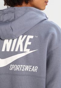 Grå hoodie med en stor vit "NIKE"-logotyp och texten "SPORTSWEAR". Tyget är mjukt, har en standardpassform och en dragsko i huven.