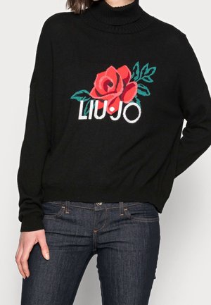 Osoba ubrane w czarny golf z grafiką czerwonej róży i zielonych liści oraz napisem „LIU•JO”, w połączeniu z ciemnoniebieskimi dżinsami.