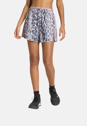 Mujer con pantalones cortos deportivos gris y negro con estampado de leopardo y rayas rosas, calcetines negros y zapatillas deportivas negras, de pie sobre fondo blanco.