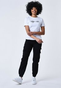 Maglietta bianca con scritte e logo neri, abbinata a pantaloni cargo neri con tasche e sneakers bianche. Design casual, aderente.
