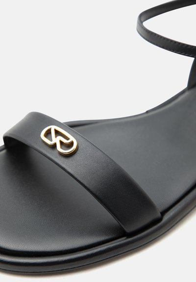 Μαύρο σανδάλι με ανοιχτά δάχτυλα, λεπτές λουρίδες και μικρό χρυσό διακοσμητικό λογότυπο στη μπροστινή λουρίδα, λεία σόλα, πλάγια κεκλιμένη όψη.