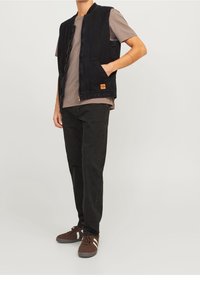 Czarna dżinsowa kamizelka z zamkiem, dwiema bocznymi kieszeniami i małą pomarańczową naszywką, w połączeniu z szarym T-shirtem i czarnymi spodniami.