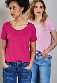 Dos modelos vistiendo camisetas de manga corta: una en rosa brillante y la otra en rosa claro. Ambas combinan con jeans de mezclilla.