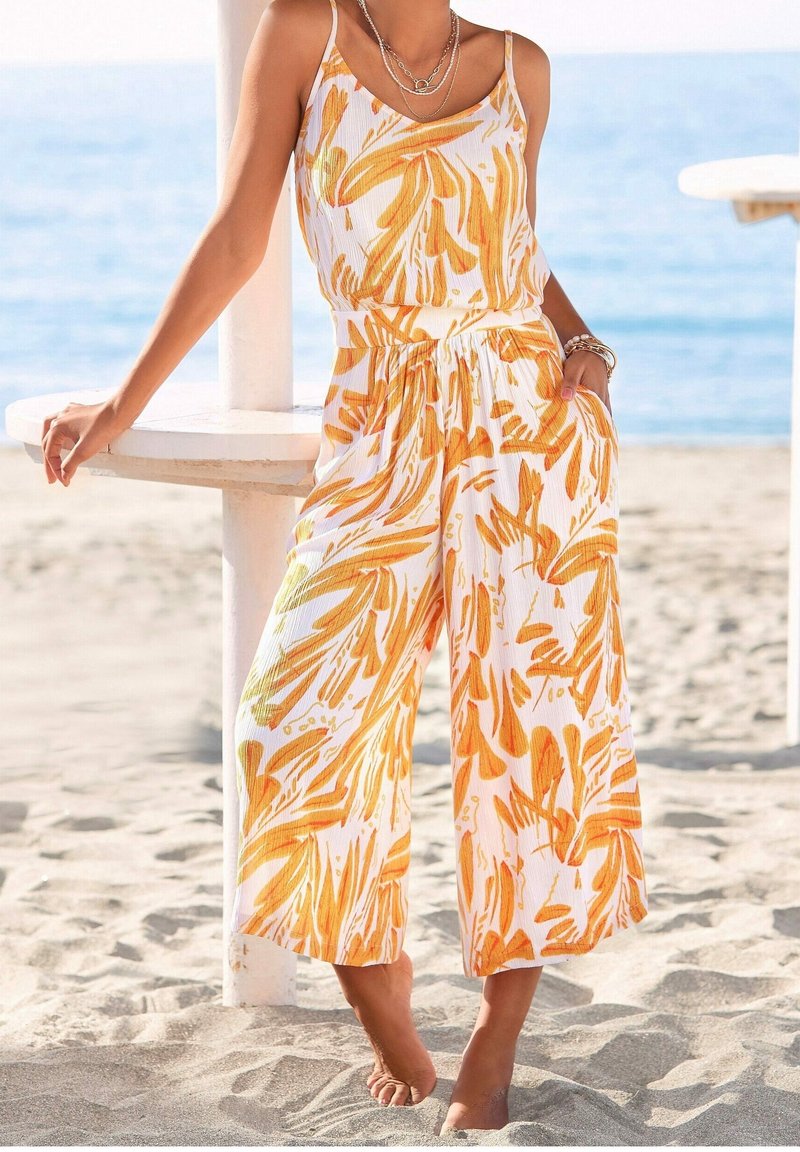 Femme en tailleur-pantalon sans manches à jambes larges avec un motif de feuilles orange, debout pieds nus sur une plage de sable près d’un poteau blanc.