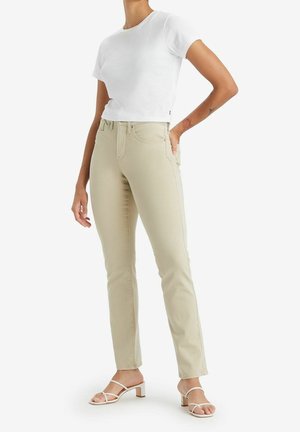 Frau trägt eng anliegende beige Hose, weißes kurzärmeliges Hemd und weiße Sandalen mit Absatz, steht mit einer Hand in der Tasche.