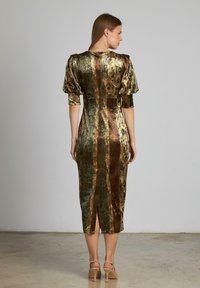 Donna che indossa un abito midi in velluto verde e marrone con maniche a sbuffo e uno spacco sul retro, in piedi su un pavimento in cemento contro un muro semplice.