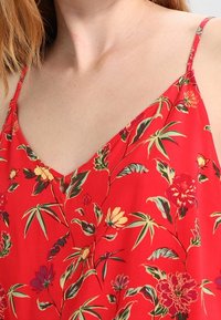 Rød blomstret cami-top med tynde stropper, V-hals og en lille nøglehul detalje i midten. Materialet har en glat, let tekstur.