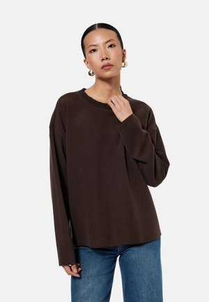 Mujer con cabello oscuro recogido en un moño bajo, lleva pendientes de aro dorados, un suéter suelto marrón oscuro y vaqueros azules sobre un fondo liso y claro.