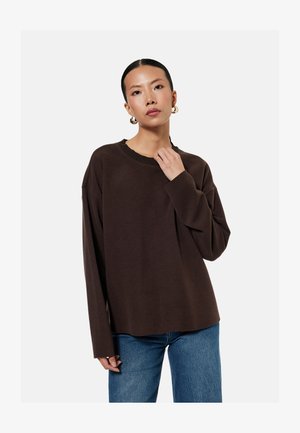 Kobieta o ciemnych włosach związanych w niski kok nosi złote kolczyki koła, luźny ciemnobrązowy sweter i niebieskie dżinsy na jednolitym jasnym tle.