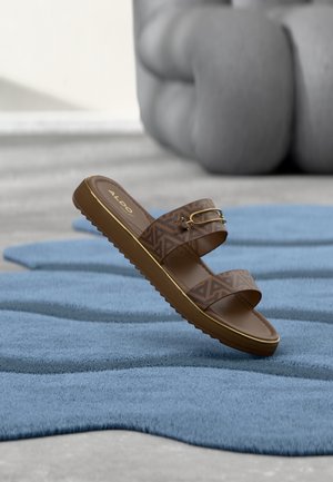 Sandale mule marron Aldo avec des brides à motifs et une boucle dorée, reposant sur un tapis bleu texturé avec un fond gris abstrait.