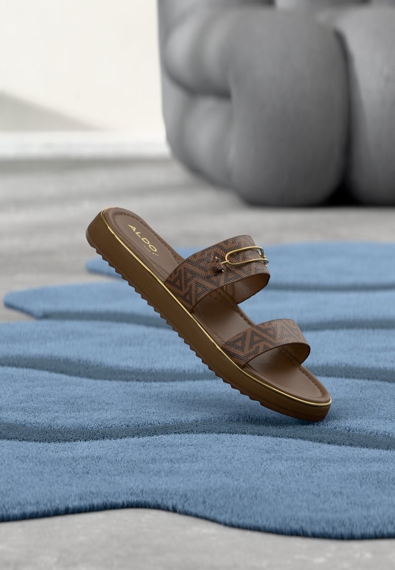 Sandale mule marron Aldo avec des brides à motifs et une boucle dorée, reposant sur un tapis bleu texturé avec un fond gris abstrait.