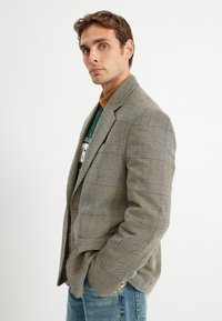 Grau kariertes Blazer aus strukturiertem Stoff; hat breite Revers, strukturierte Schultern und zwei Knöpfe. Getragen über einem grünen Hemd.