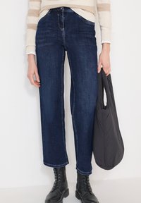 Personne portant un jean bleu foncé, des bottes noires à lacets, un pull beige rayé, tenant un sac matelassé noir sur un fond blanc.