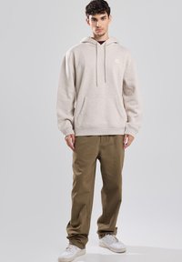Beige Kapuzenpullover mit einer Vordertasche und Kordeln, kombiniert mit olivgrünen Hosen und weißen Turnschuhen. Minimales Design, weiche Textur.