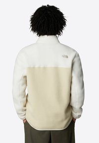 Fleecejakke i krem og beige med høy krage, som har en jevn tekstur og subtil logo detalj på øvre rygg.