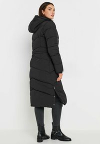 Cappotto imbottito nero con cappuccio, caratterizzato da una silhouette lunga, design a zigzag e dettaglio con bottoni a pressione all'orlo. Il modello indossa stivali alla caviglia.