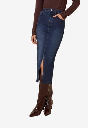 Kvinde iført en mørkeblå denim midi-nederdel med frontslids, kombineret med brune knælange støvler med hæl og en brun langærmet top.