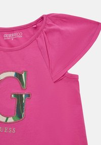 Camicia in cotone organico rosa con maniche a sbuffo corte, caratterizzata da un logo "G" decorativo in camo verde e blu e dettagli ricamati.