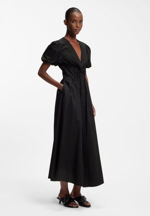 Femme debout portant une longue robe noire avec des manches bouffantes, des boutons à l'avant, un profond décolleté en V, des poches et des sandales à talons noires avec des boucles.