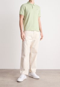 Homme portant un polo à manches courtes vert clair, un pantalon beige à jambes larges et des baskets blanches, se tenant sur un sol gris contre un mur blanc.