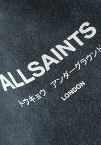 Tummanharmaa kangas, jossa valkoisella tekstillä lukee "ALLSAINTS" ja lisäksi merkkejä japaniksi. Kangas on pehmeä ja suunnittelultaan rento.