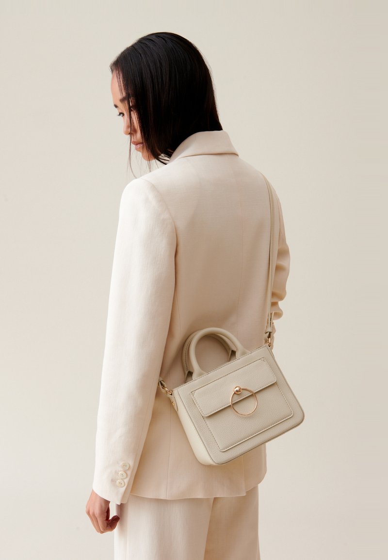 Sac à main structuré beige avec une poignée supérieure, une forme rectangulaire et un accent en métal circulaire sur le devant. Porté avec un costume de couleur claire.