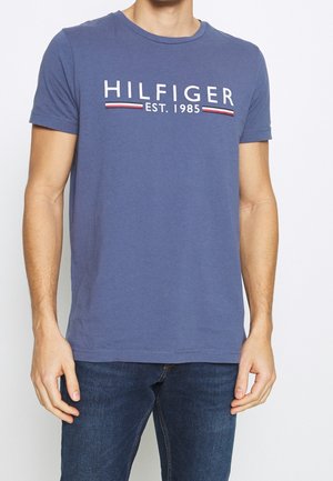 Homme portant un T-shirt bleu à manches courtes avec le texte « HILFIGER EST. 1985 » et des bandes rouges, blanches et noires sur la poitrine.