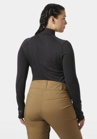 Žena s copánky, oblečená v černém topu s dlouhým rukávem a hnědých kalhotách s vysokým pasem, zobrazená zezadu na jednobarevném pozadí.