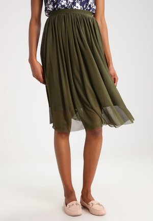 Femme portant une jupe midi plissée vert olive, un haut à motifs floraux et des mocassins rose clair, debout les mains détendues le long du corps.