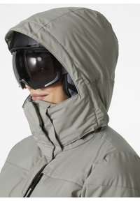 Helle graue isolierte Jacke mit Kapuze und hohem Kragen, mit Reißverschluss und Stofftextur, kombiniert mit schwarzen Skibrillen.