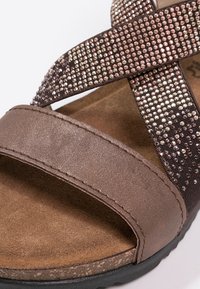 Sandale marron avec un design de lanières croisées, présentant des accents métalliques et des strass intégrés. Semelle intérieure en suède doux et semelle en caoutchouc texturé.