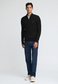 Czarny sweter w splot bądź wszerny z kołnierzem na ćwierć zamka, zestawiony z niebieskimi dżinsami i beżowymi sneakersami, stojący na tle jasnym.