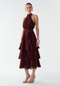 Burgundi, be rankovėmis su aukštu kaklu, prigludęs liemuo ir sluoksniuotas volanų sijonas. Pakuotėje yra suderintas diržas juosmeniui ir permatomos medžiagos sluoksniai.