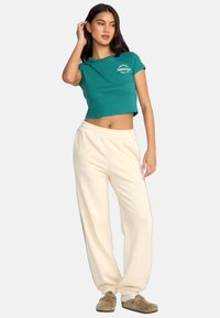 Przycięta, turkusowa bawełniana koszulka z białym logo, w połączeniu z luźnymi, kremowymi spodniami dresowymi i brązowymi butami wsuwanymi.