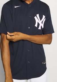 Laivastonsininen Nike-pelipaita, jossa on lyhyet hihat, napillinen edusta ja vaaleanpunainen New York Yankees -logo. Paidassa on resorikuviointi ja helman kohdalla oleva lappu.