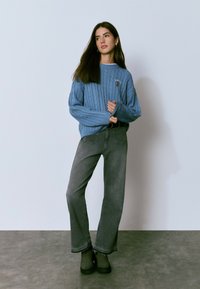 Junge Frau steht und trägt einen blauen Zopfmusterpullover, graue weite Jeans und schwarze klobige Stiefel vor einem einfarbigen grauen Hintergrund.