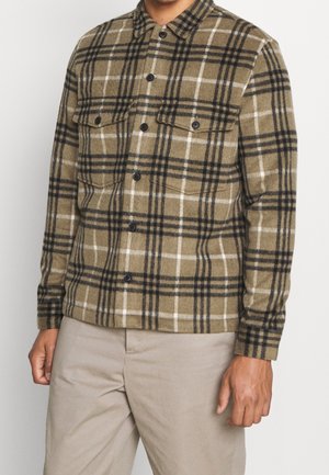 Mand iført en brun og sort ternet skjorte med brystlommer og beige bukser, stående mod en ensfarvet hvid baggrund.
