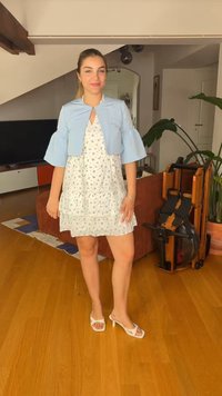 Jeune femme debout à l'intérieur, portant une robe blanche à fleurs, une veste courte bleu clair et des sandales blanches à talons dans un salon.