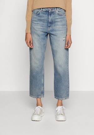 Jasnoniebieskie jeansy z wysokim stanem i prostą nogawką z delikatnym przetarciem na prawej udzie, połączone z beżowym swetrem i białymi trampkami na jednolitym tle.