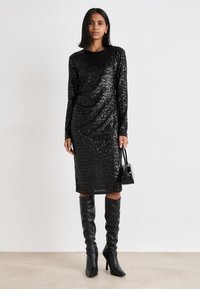 Vestido preto com lantejoulas, com mangas longas, corte até ao joelho e design drapeado; combinado com botas altas de couro pretas e uma pequena bolsa.