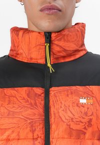 Veste matelassée avec une partie supérieure à motif floral orange et une partie inférieure noire. Elle dispose d'un col haut, d'une fermeture éclair et d'un cordon de serrage jaune.