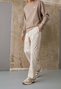 Beige sweatshirt med vit logga, ljusa byxor med subtilt märkesdetalj, och beige löparskor med grå accenter på en strukturerad bakgrund.