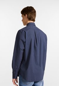 Camicia blu navy con bottoni, caratterizzata da un motivo sottile, maniche lunghe e un design con piega posteriore. Realizzata in un tessuto leggero e morbido.