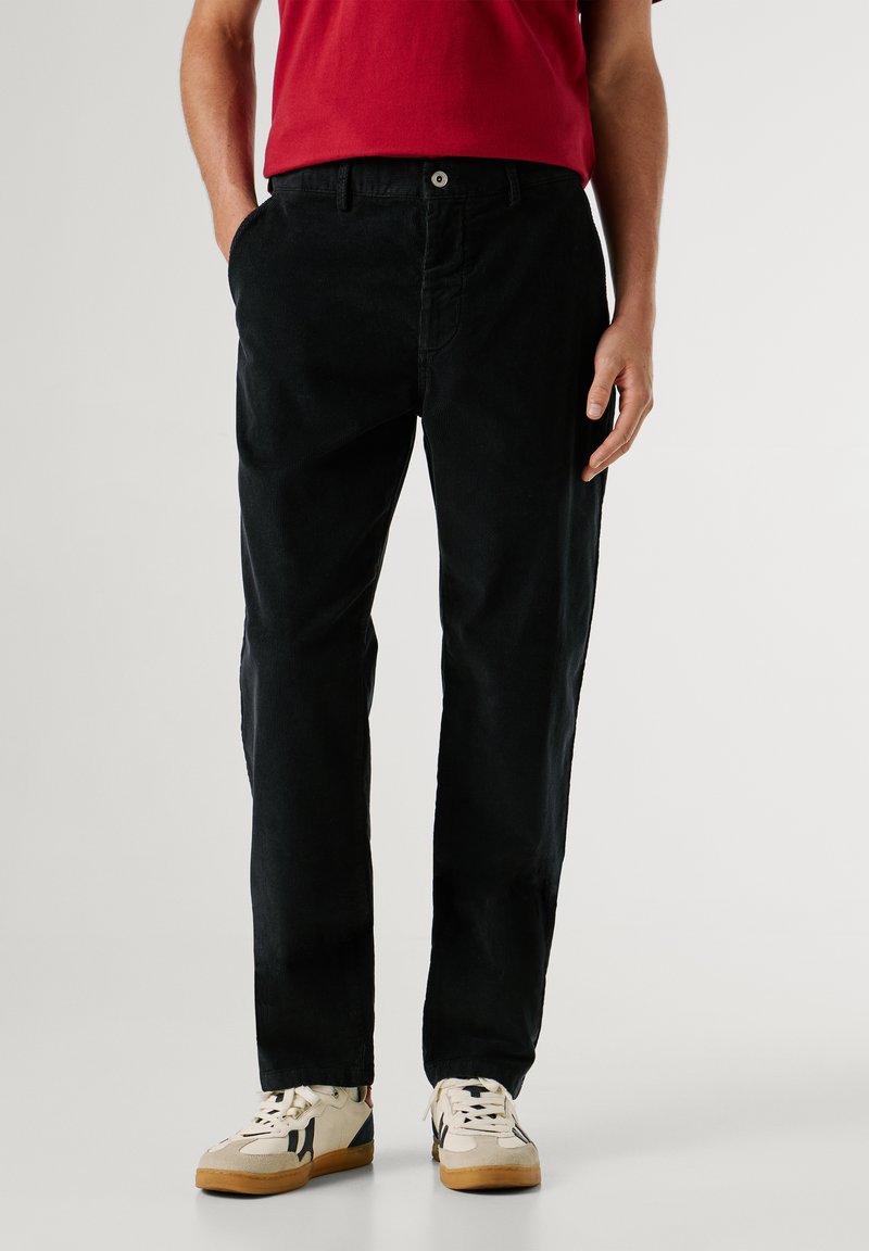 Zwarte corduroy broek met een relaxte pasvorm, voorzien van een knoopsluiting en twee zijzakken, gecombineerd met beige en gemusterde sneakers.