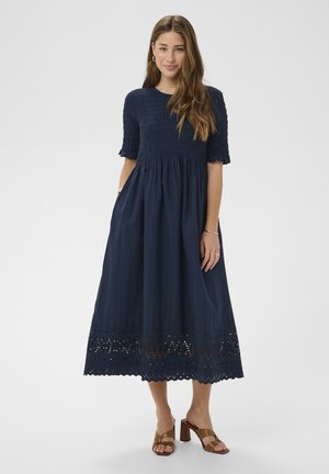 Woman wearing a navy blue midi dress with short sleeves, eyelet lace hem, and brown heeled sandals, standing against a plain background.
