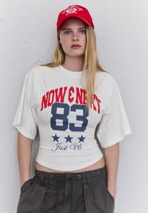 Mladá žena s dlhými blond vlasmi nesúca červenú šiltovku, biele krátke tričko s textom "NOW & NEXT 83" a tmavé voľné nohavice, ruky v vreckách.